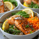 Served salmon bowl with salmon, grains, avocado, vegetables, sesame seeds, and lemon on a dinner table.